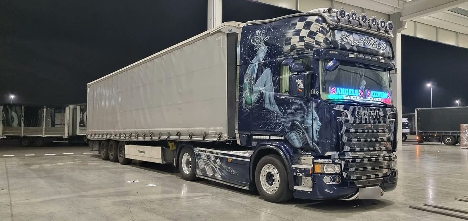 Uno Scania blu della flotta Atlante con rimorchio centinato, fermo in piazzale di notte. Sul parasole la scritta Team Atlante.